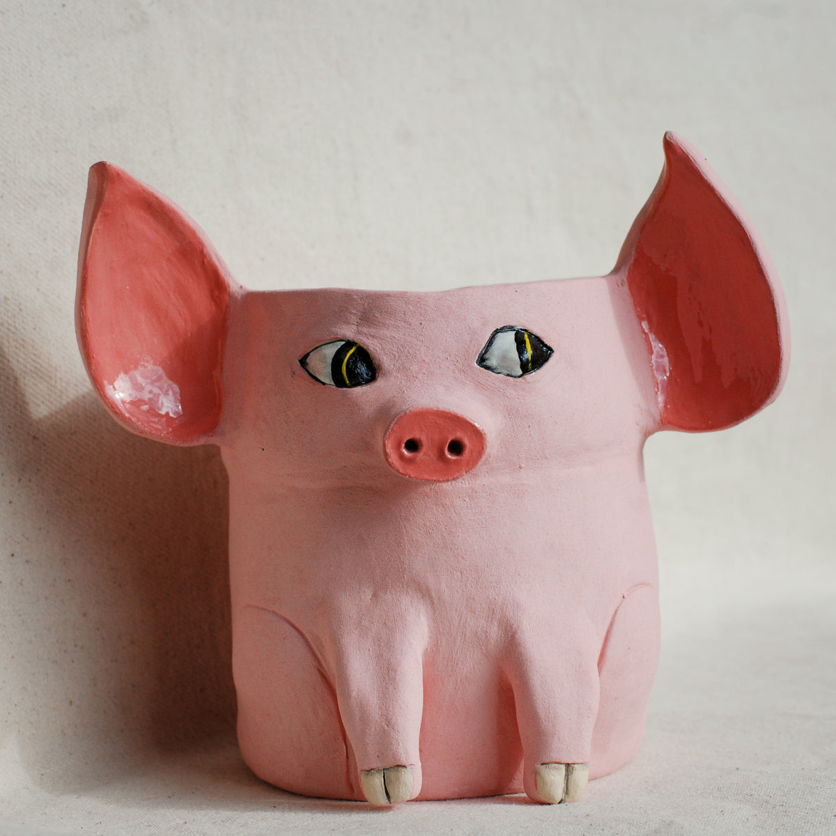 A pink pig-shaped planter with large pointed ears, painted eyes, and small hooves, photographed against a plain light beige wall.