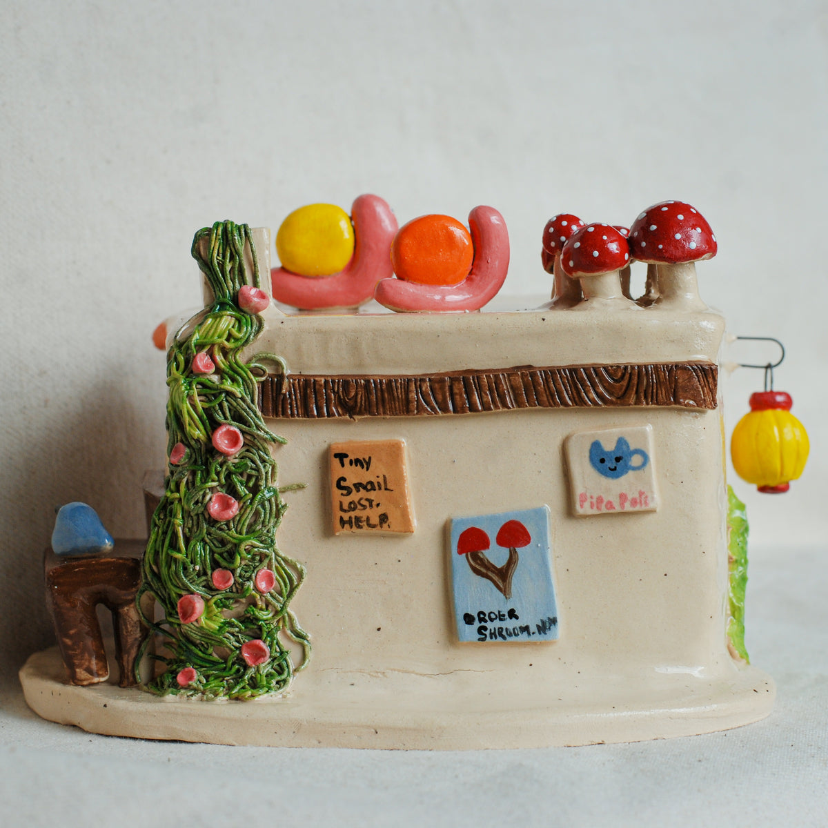 A handmade ceramic miniature house with whimsical details, including a green vine-covered chimney, red and white spotted mushrooms on the roof, colorful tiles, and small decorative elements, set against a plain light background.