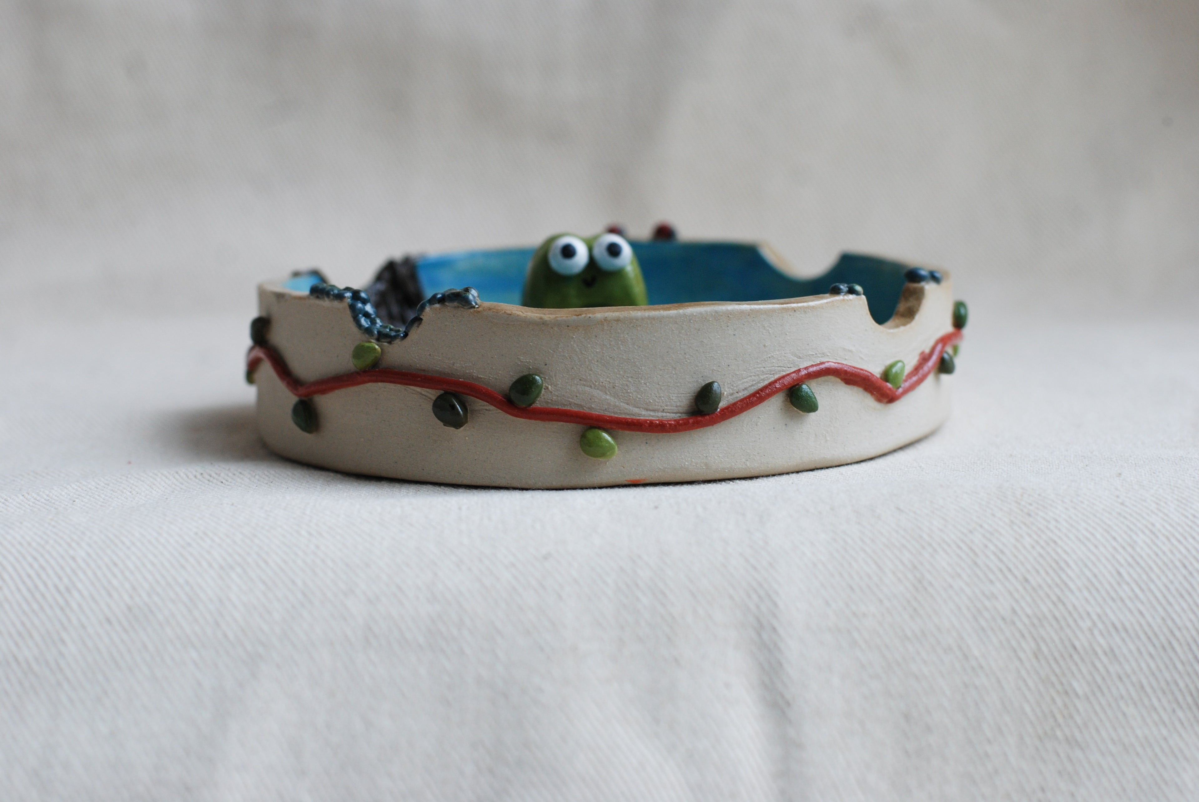 Ceramic ashtray made by pipa pots features a cute frog poping out with decorative elements on a white background