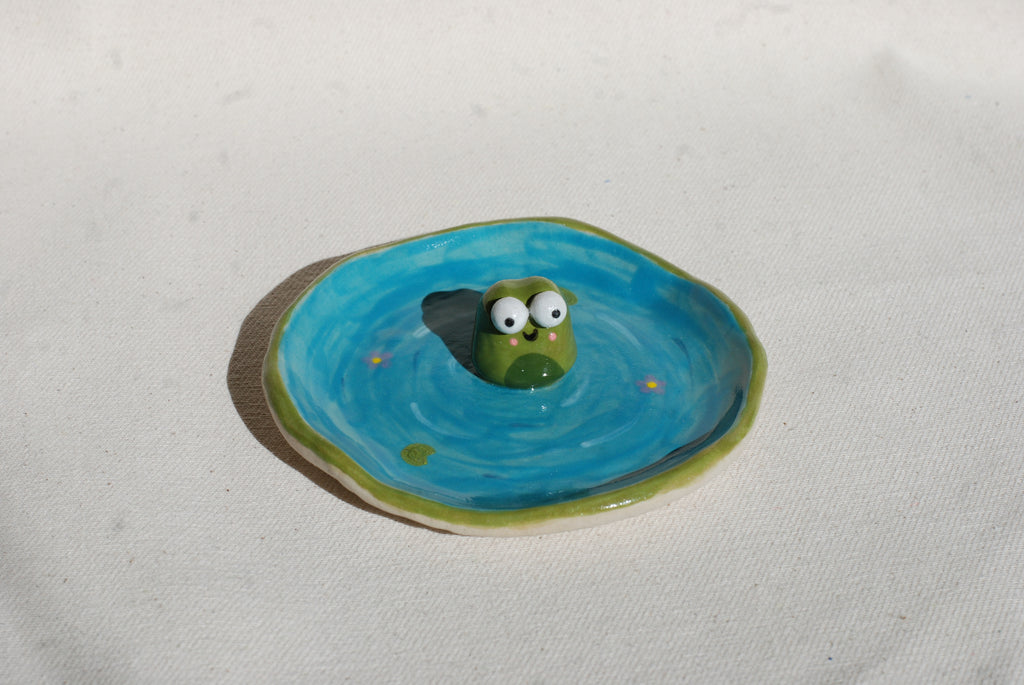 Small ceramic frogfigurine on a blue circular base with a white background made by pipa pots in the netherlands
