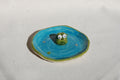 Small ceramic frogfigurine on a blue circular base with a white background made by pipa pots in the netherlands