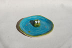 Small ceramic frogfigurine on a blue circular base with a white background made by pipa pots in the netherlands
