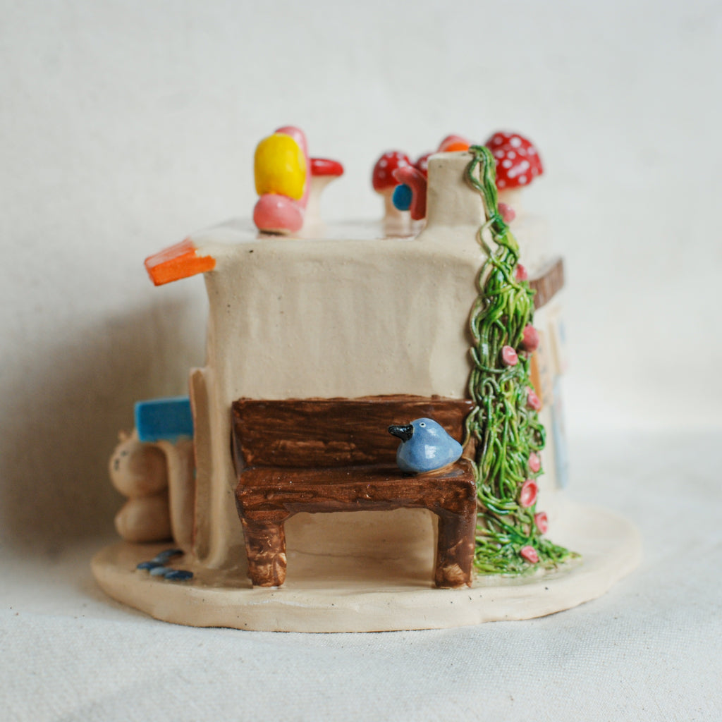 A miniature ceramic fairy house with a small wooden bench, blue bird, red and white mushrooms, and green vine details, set against a plain light background.