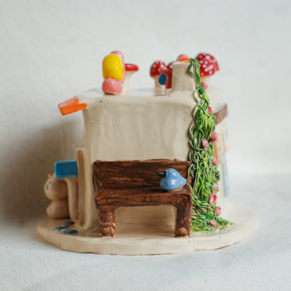 A miniature ceramic fairy house with a small wooden bench, blue bird, red and white mushrooms, and green vine details, set against a plain light background.