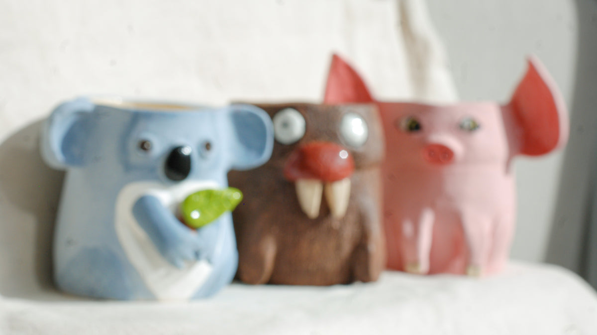 Three small animal-shaped ceramic containers or pots in blue, brown, and pink, arranged side by side on a white surface against a light wall.