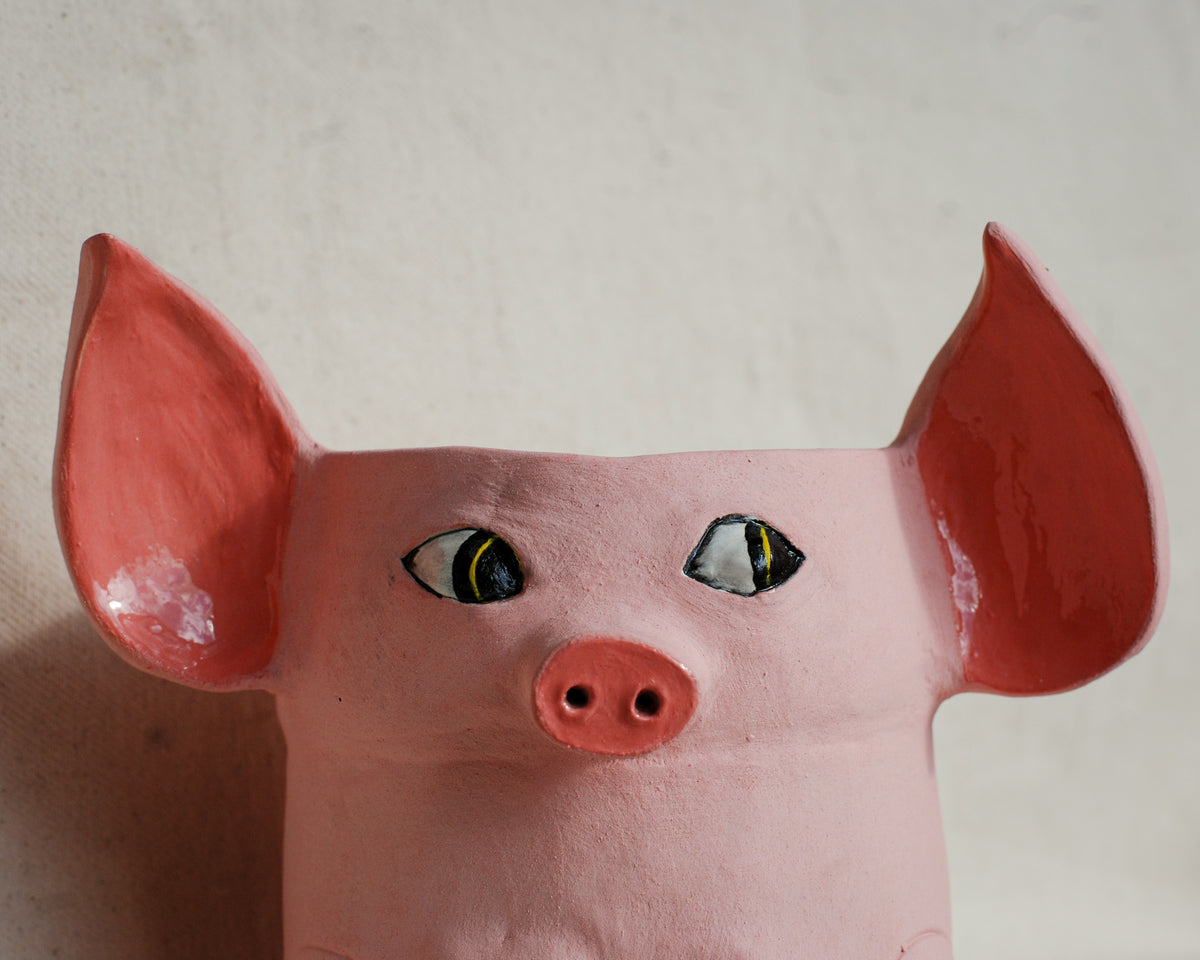 A pink pig-shaped planter or decorative pot with painted red ears, black and yellow eyes, and a red snout, photographed against a plain light beige wall.
