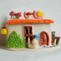 A whimsical ceramic fairy house figurine with a beige body, orange roof, green textured bushes, red-and-white mushrooms, a small cat, a mailer box, and hanging lanterns, photographed against a plain light background.
