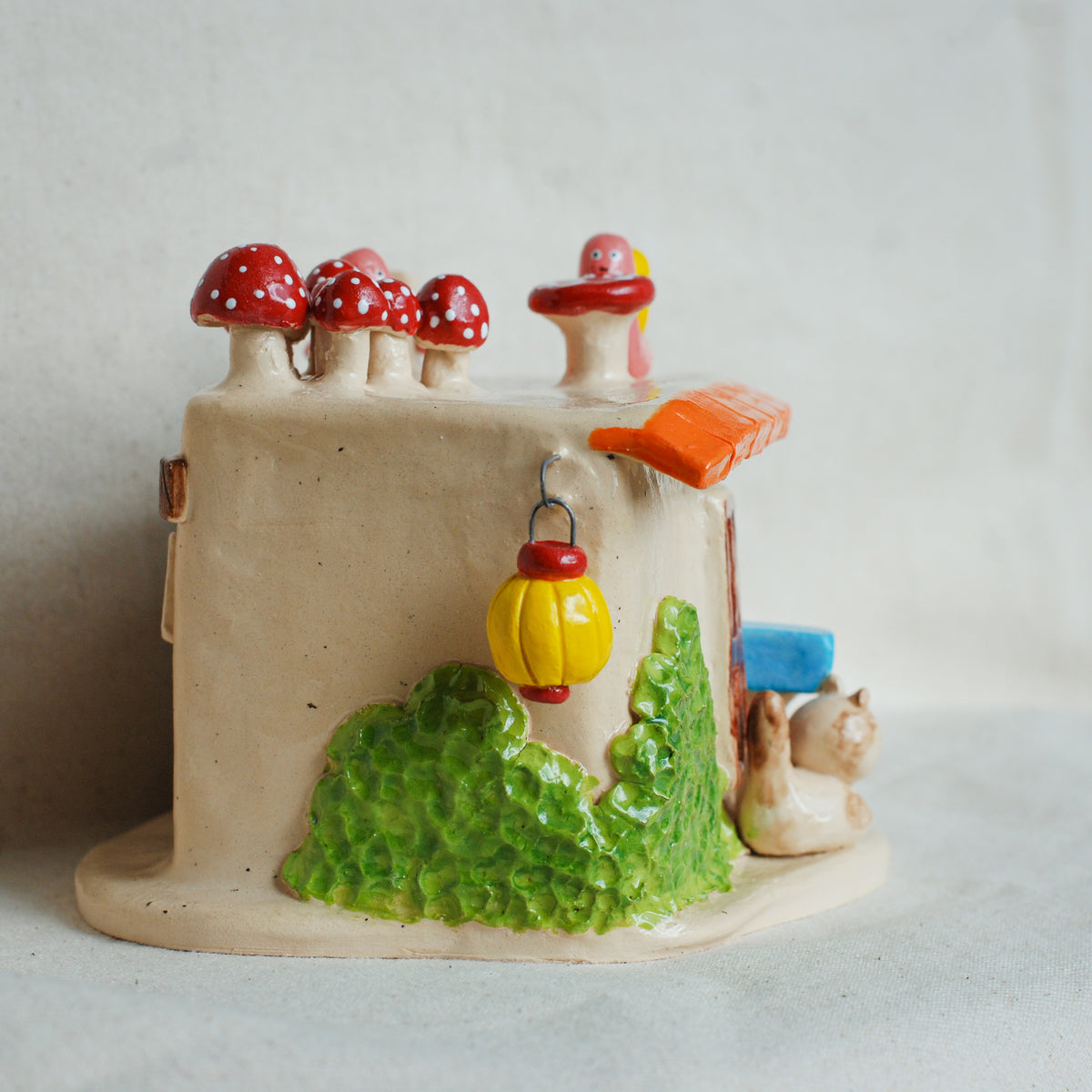 A small ceramic fairy house figurine with a beige body, red and white mushrooms on top, a green textured base, a yellow hanging lantern, and a small cat figure at the side, photographed against a light neutral background.